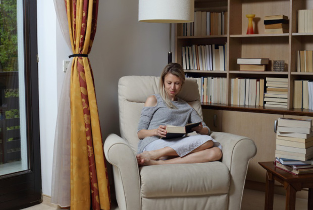 Young woman wearing cozy sweater,sitting in comfortable armchair, enjoying reading a book at home library